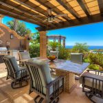 Outdoor dining area of a luxury villa with a stone table, cushioned chairs, barbecue grill, and ocean view surrounded by lush vegetation.
